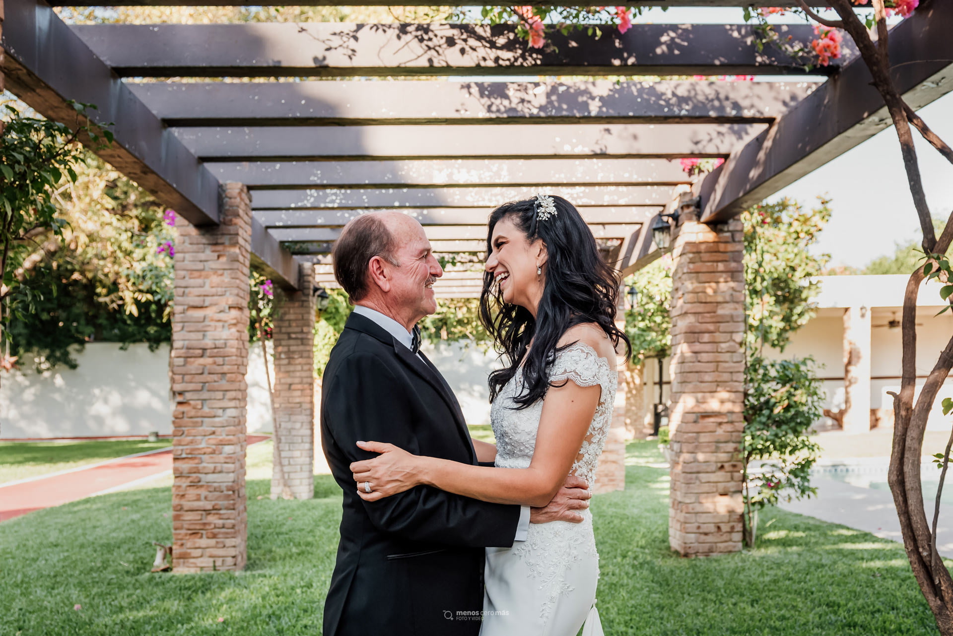 Alejandra y Chuy, radiantes de felicidad en su foto formal de boda, se abrazan frente a frente en un jardín en San Pedro Garza García al fondo. La ceremonia religiosa se realizo en Parroquia San Agustín.