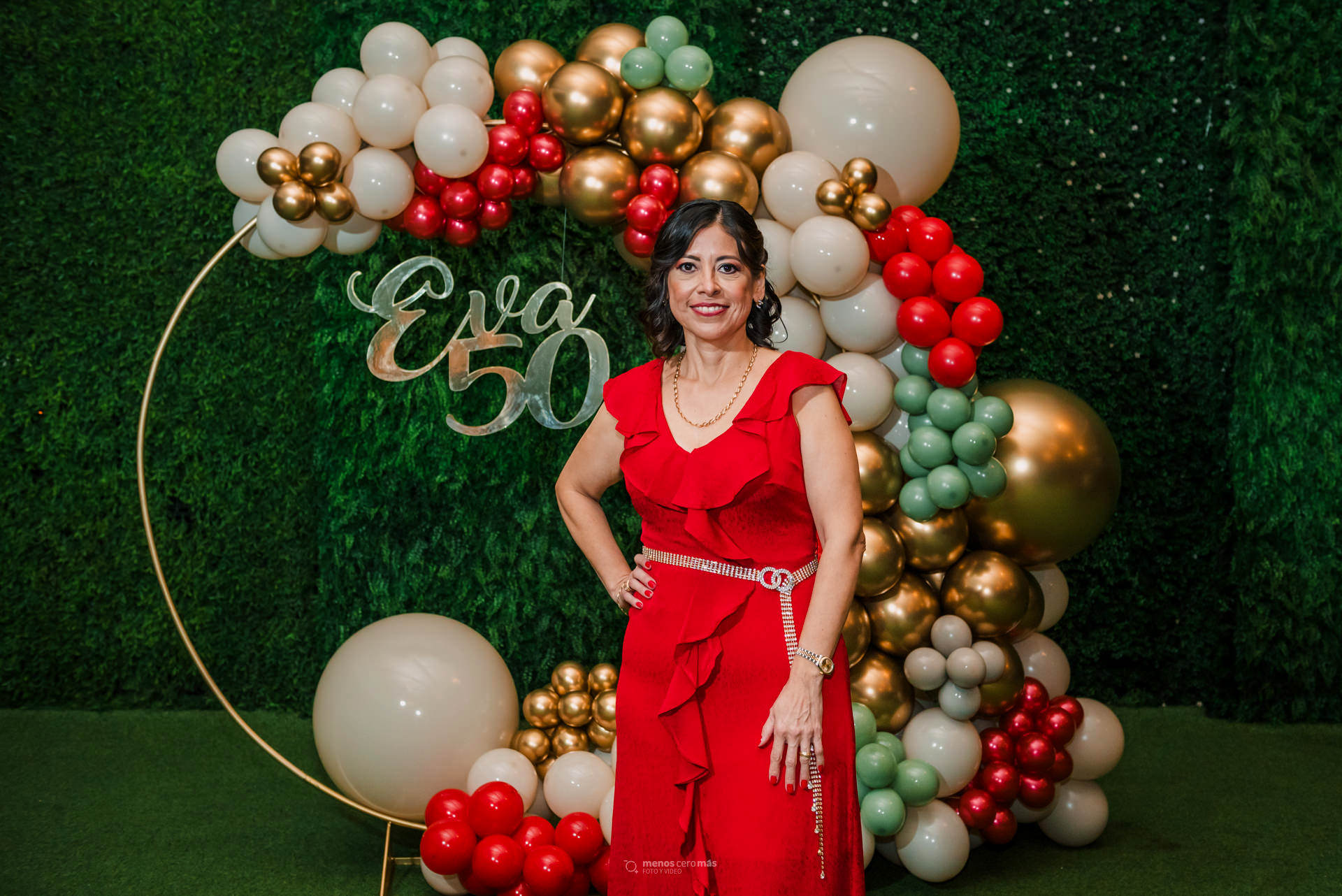 Eva irradia felicidad en su 50 aniversario, celebrado en Las Lomas Eventos con una elegante decoración en tonos rojos, beige y verdes. La imagen, capturada por Menosceromás Fotografía, muestra a la homenajeada sonriente frente a un letrero que dice "Eva 50"
