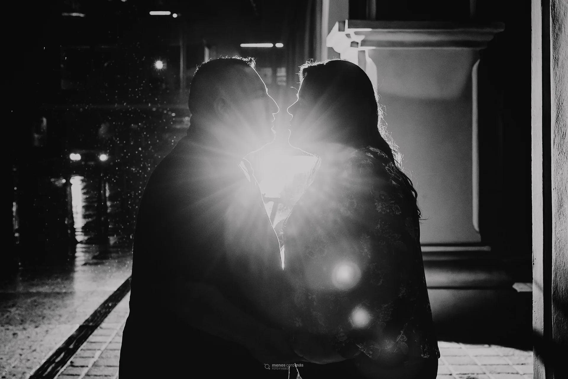Fotografía de Ana y Ernesto en una galería informal de fotos, abrazados frente a una motocicleta iluminada en los pasillos exteriores del Museo Metropolitano de Monterrey, bajo la lluvia nocturna.