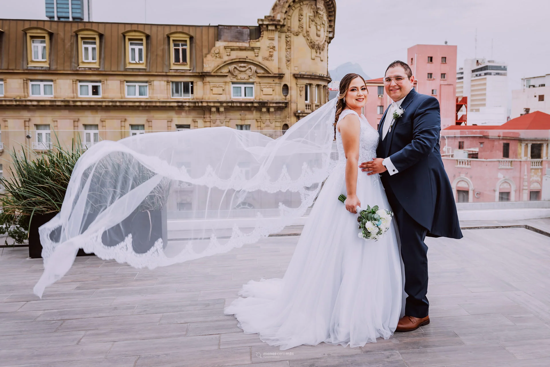 Imagen de Ana y Christian, vestidos de novios, posando frente al Hotel Ancira en Monterrey, México. La novia, con un radiante vestido blanco, abraza al novio, quien también lleva un elegante traje. Las fotos se realizaron en el mirador del Hotel Kavia. La recepción fue en Las Pampas Eventos